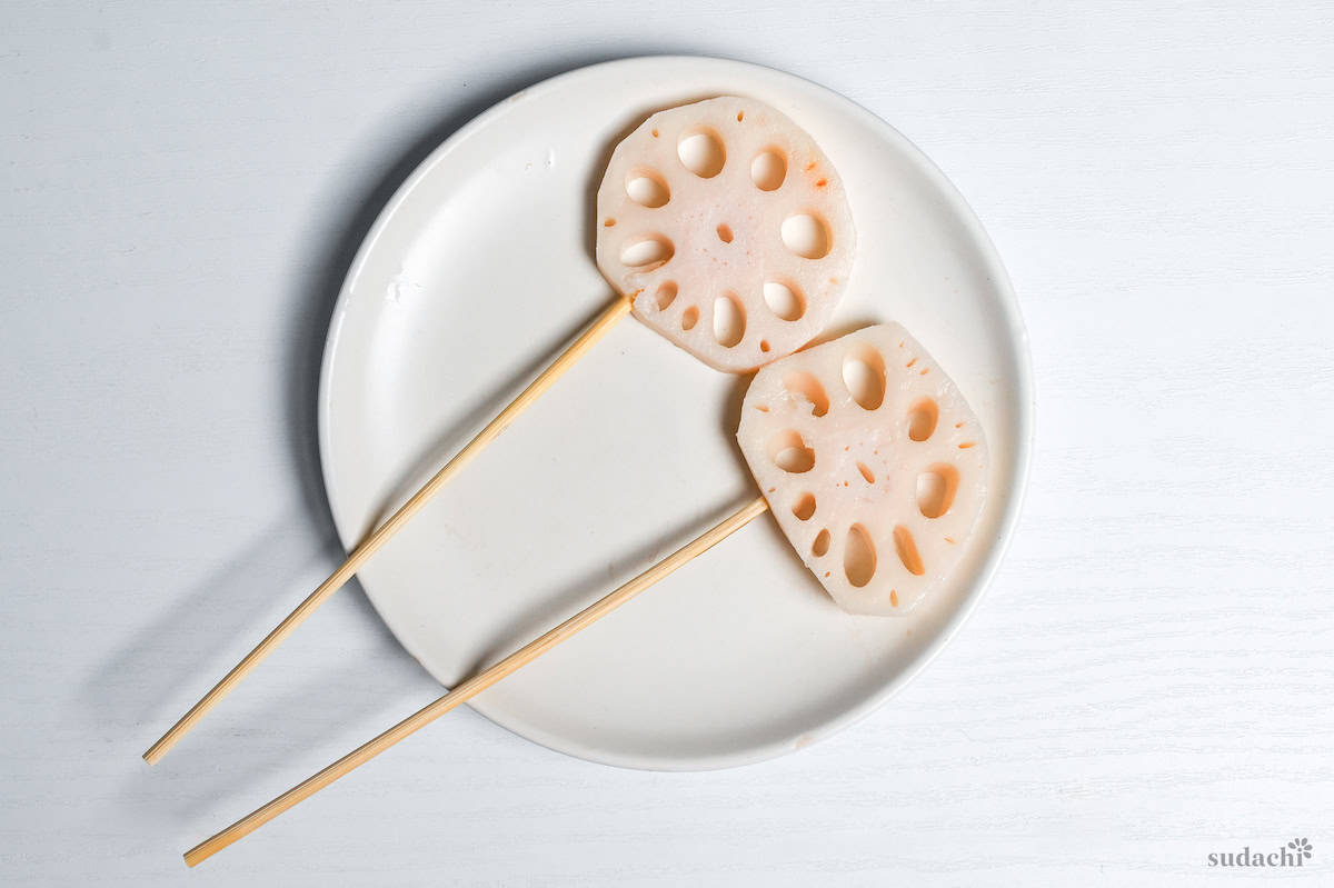 two pieces of blanched lotus root on bamboo skewers on a white plate on a white background