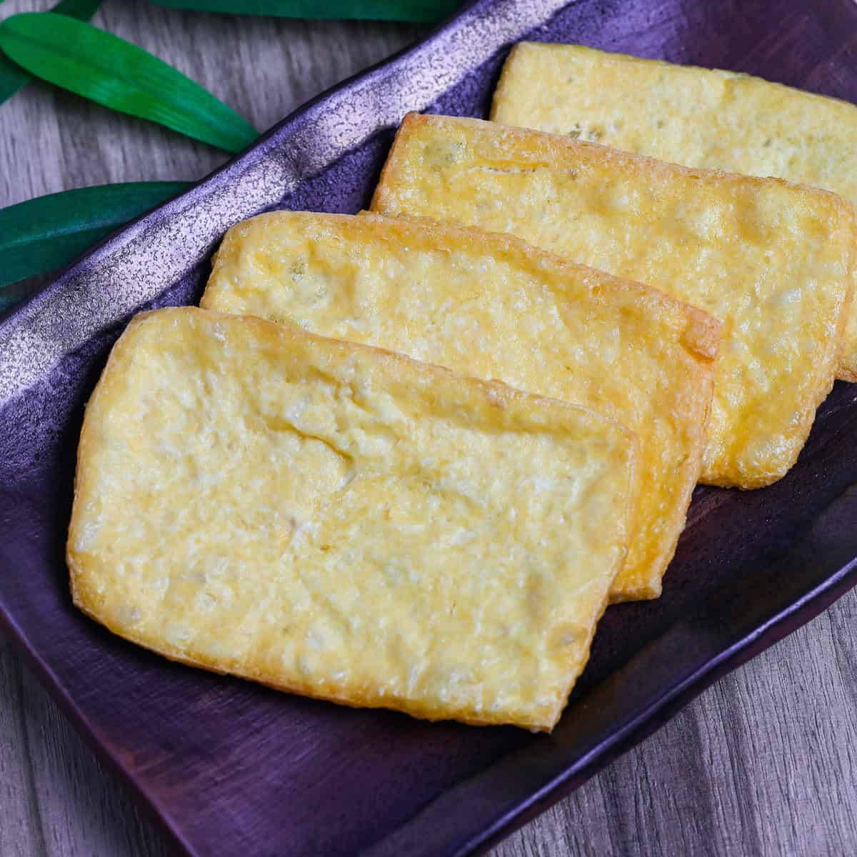 Four sheets of deep fried tofu pouches on a rectangular purplish brown plate on ashy wood surface