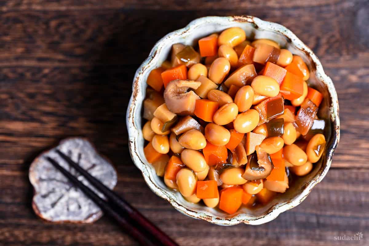 Japanese simmered soybeans (gomoku mame) in a gray textured bowl on a dark wooden surface next to wooden chopsticks on flower chopsticks rest