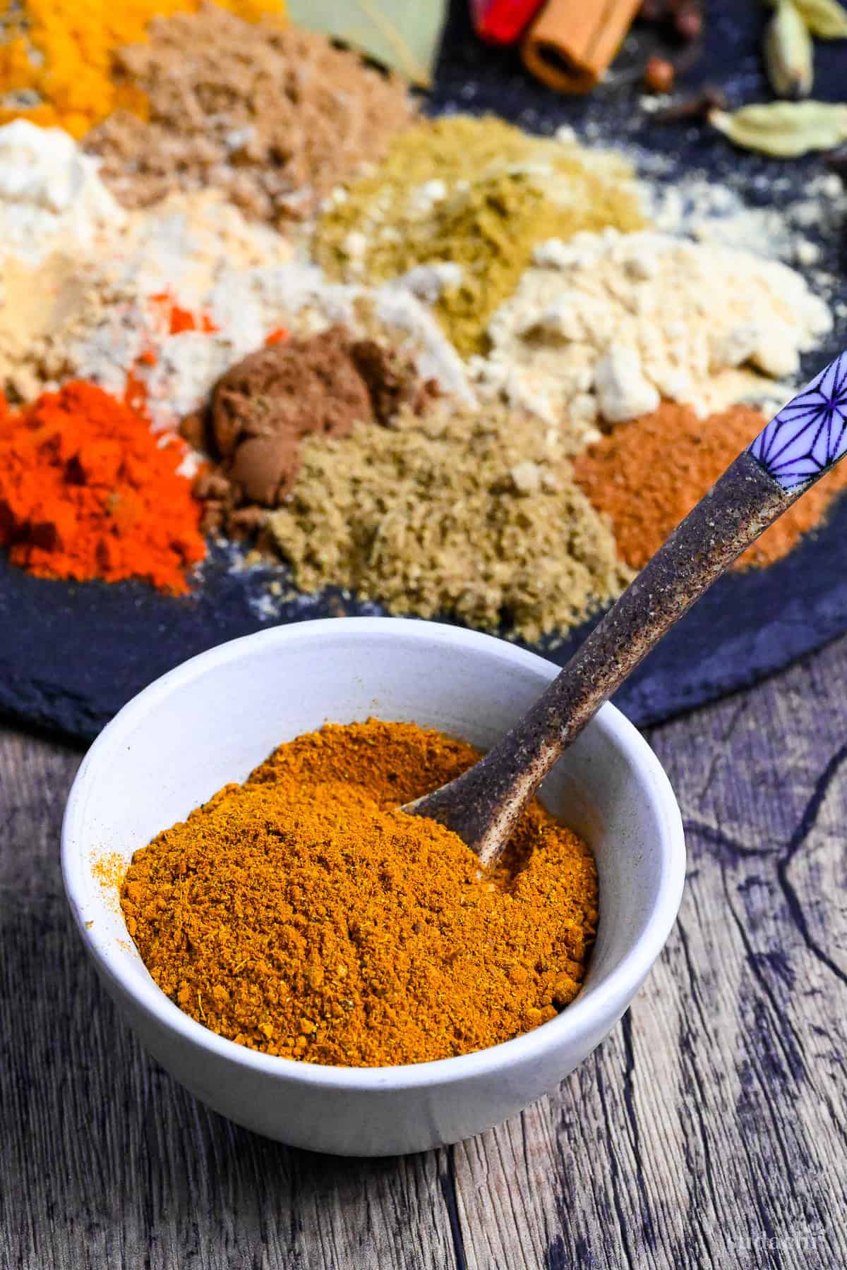 Homemade Japanese style curry powder in a small white bowl with brown ceramic spoon and a mixture of powdered spices in the background