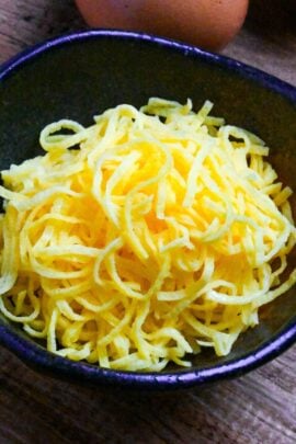 Homemade kinshi tamago (shredded egg crepe) in a black bowl with eggs in the background