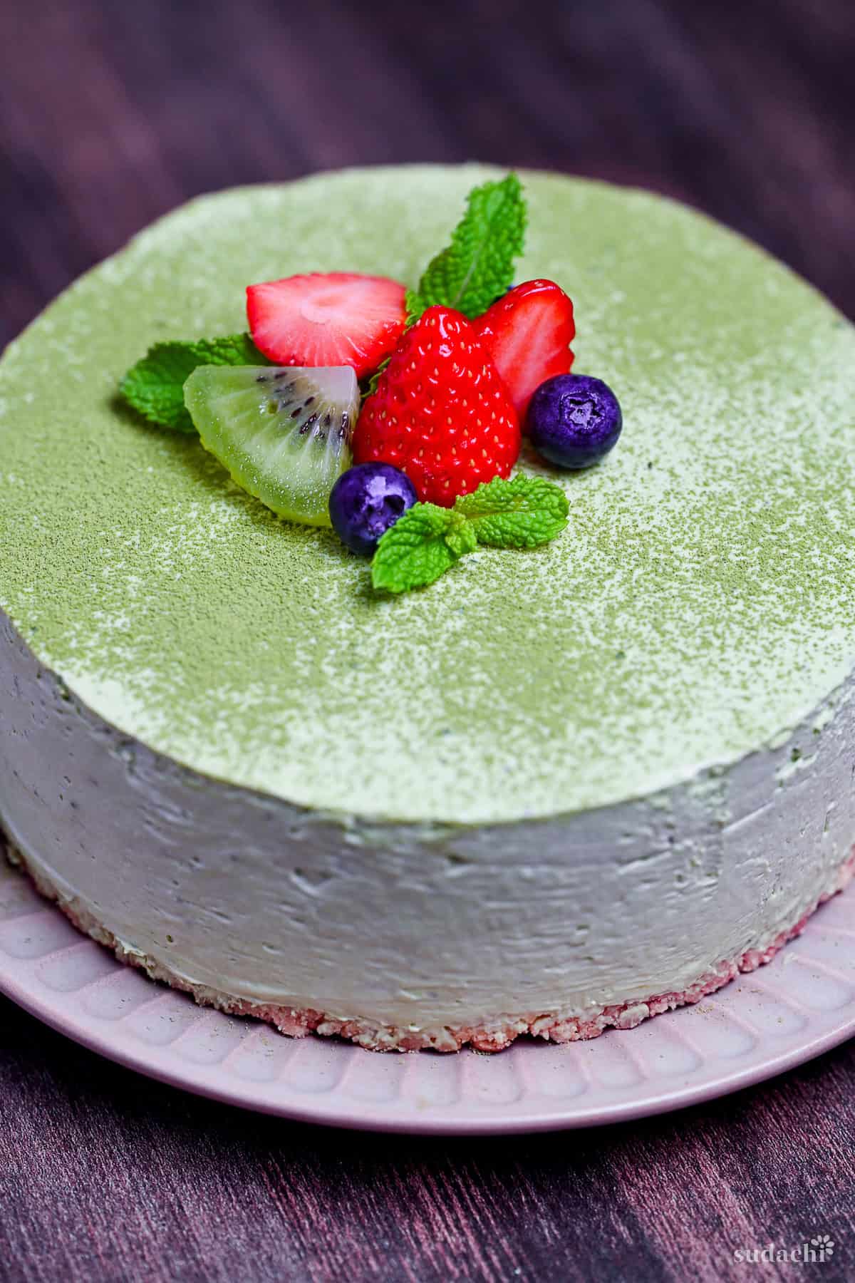 Matcha rare cheesecake on a pink plate topped with strawberries, blueberries, kiwis and mint leaves