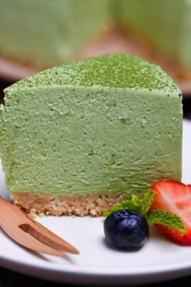 A single slice of matcha rare cheesecake on a white plate next to half a strawberry, a blueberry, and a few mint leaves
