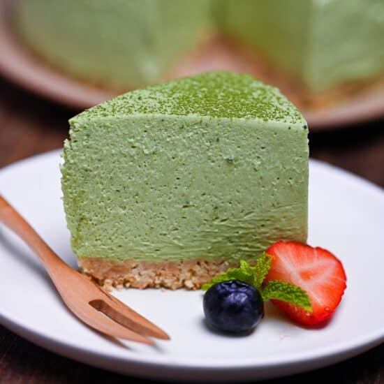 A single slice of matcha rare cheesecake on a white plate next to half a strawberry, a blueberry, and a few mint leaves