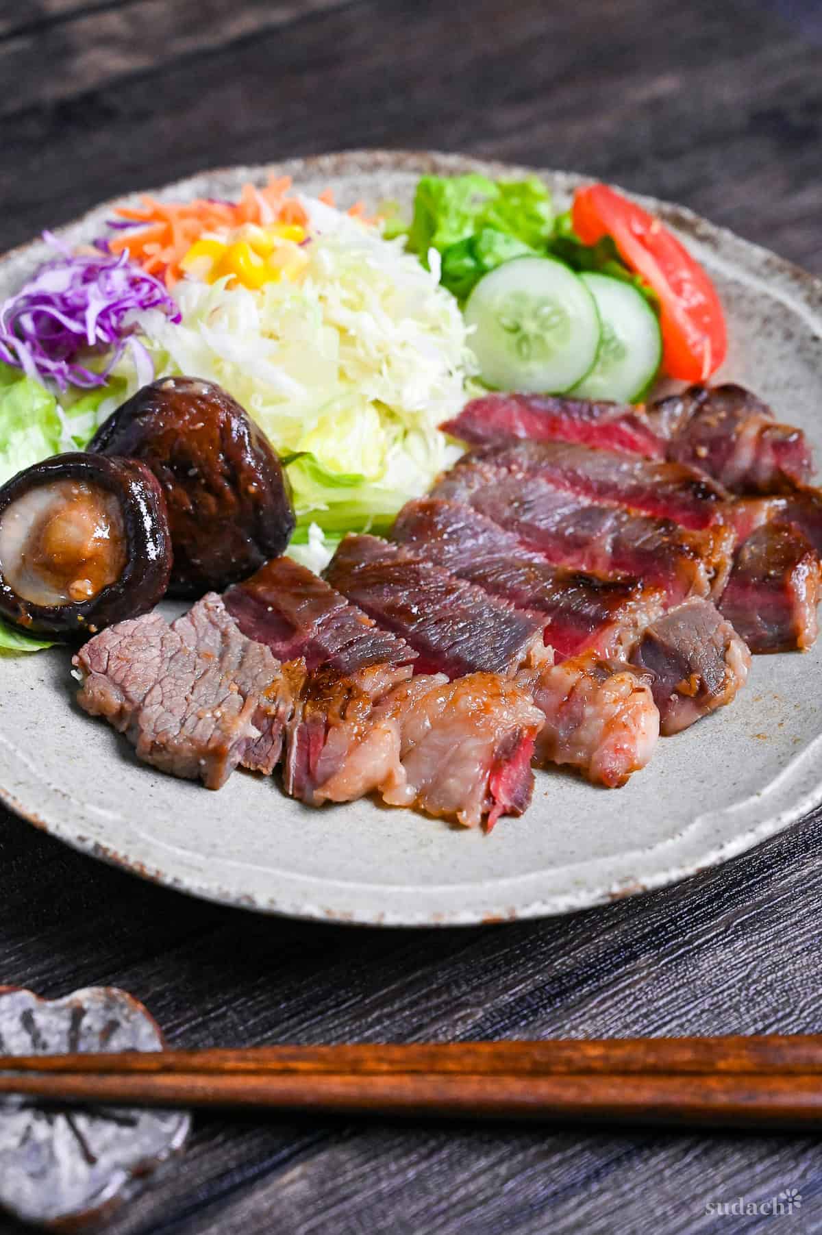 Miso marinated steak cut into slices revealing pink center on a gray plate with salad and fried shiitake mushrooms