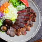 Miso marinated steak cut into slices revealing pink center on a gray plate with salad and fried shiitake mushrooms