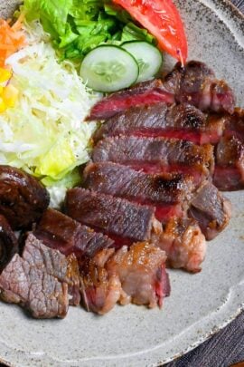 Miso marinated steak cut into slices revealing pink center on a gray plate with salad and fried shiitake mushrooms