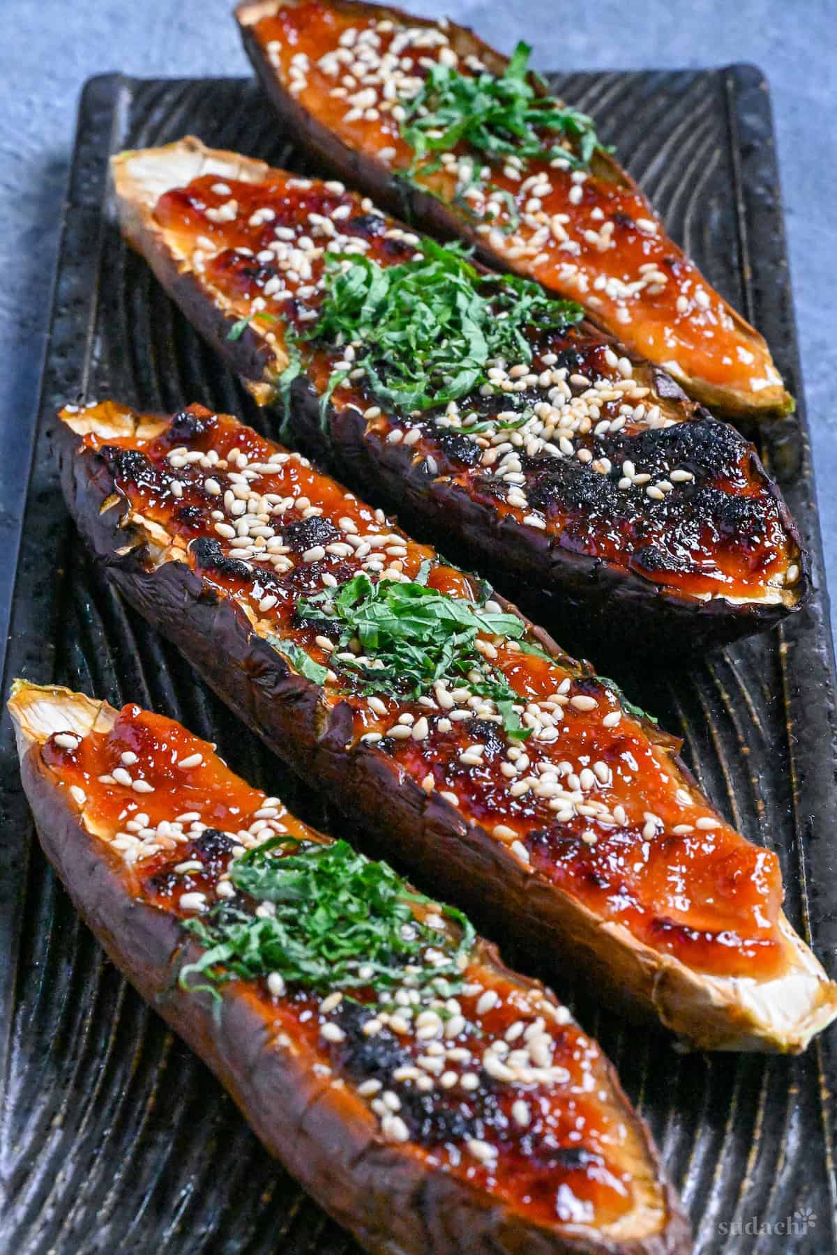 Nasu dengaku (Japanese miso-glazed eggplant) sprinkled with sesame seeds and shredded Perilla leaves on a dark textured rectangular plate