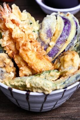 Tendon (Tempura Rice Bowl) made with shrimp and a variety of vegetables in a striped bowl on a wooden surface with homemade sauce in the background