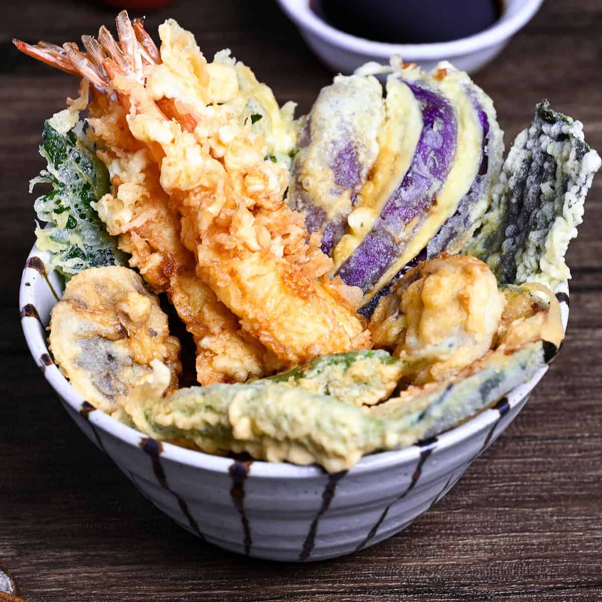 Tendon (Tempura Rice Bowl) made with shrimp and a variety of vegetables in a striped bowl on a wooden surface with homemade sauce in the background