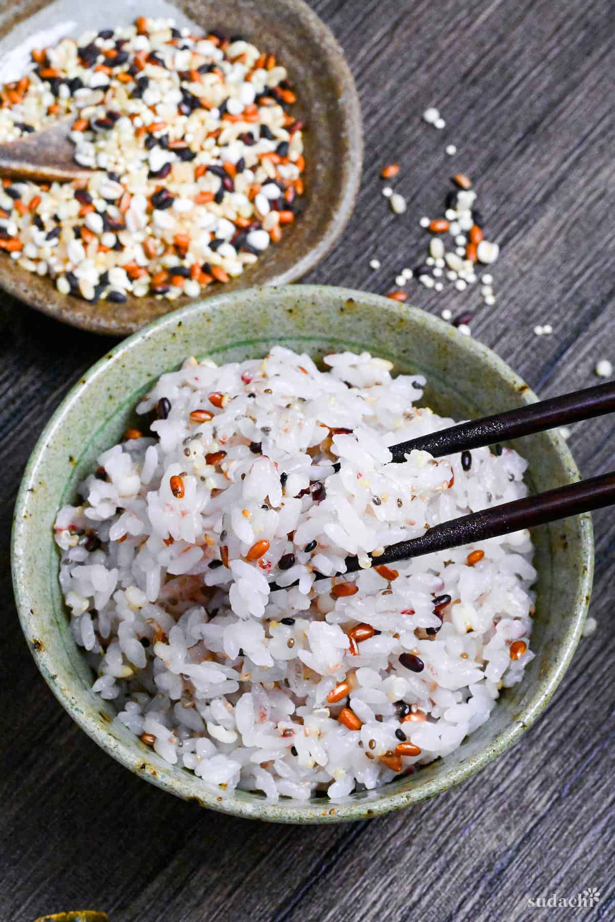 Japanese multigrain rice (zakkokumai) in a green and brown ceramic rice bowl surrounded by various grains and seeds