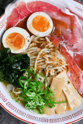 Cold ramen served in a white and red ramen bowl topped with cured ham, soft boiled eggs, wakame, chopped green onions and menma bamboo shoots on an ashy wood background