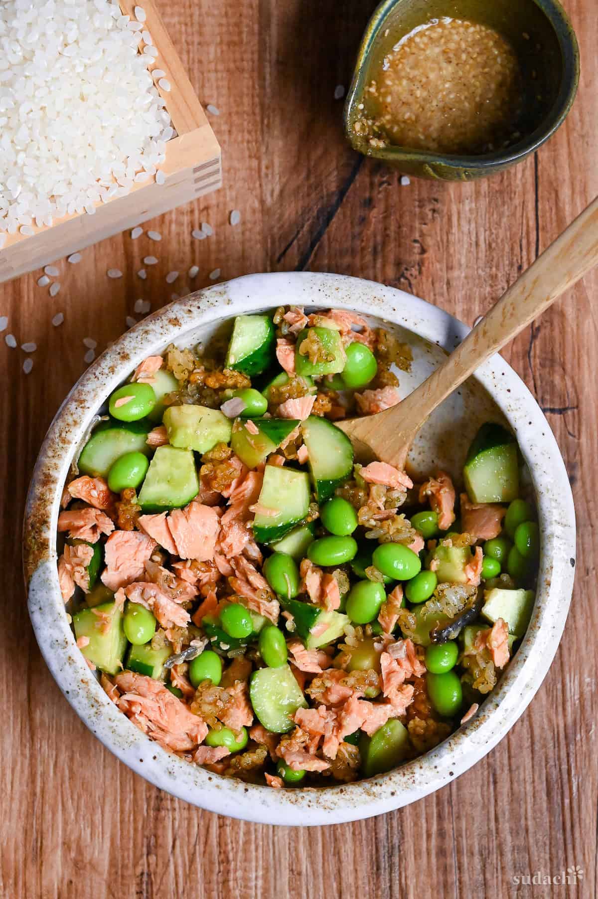 Japanese style crispy rice salad in a mottled bowl with wooden spoon next to small green jug of yuzu dressing and raw rice in a square wooden box