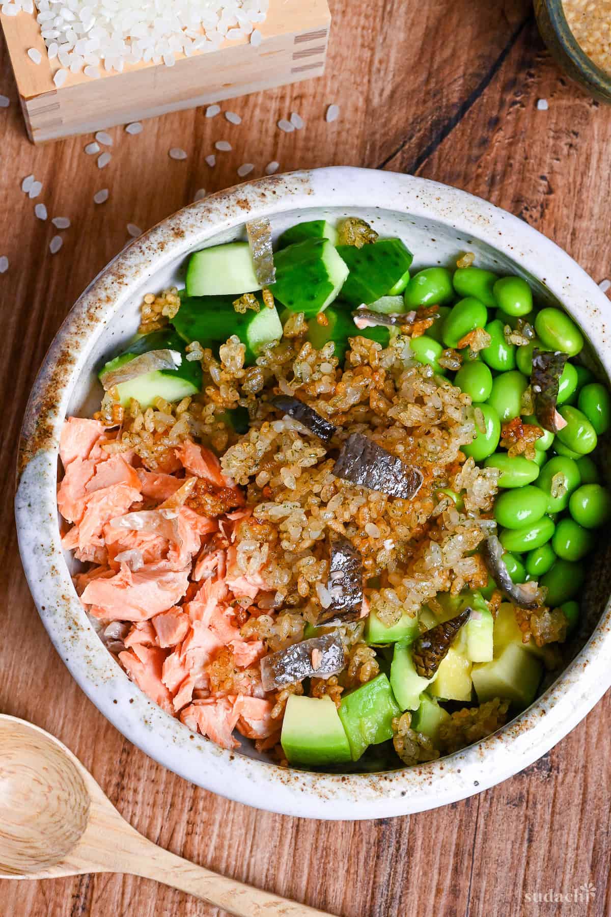 Japanese style crispy rice salad in a mottled bowl with wooden spoon next to small green jug of yuzu dressing and raw rice in a square wooden box top down view