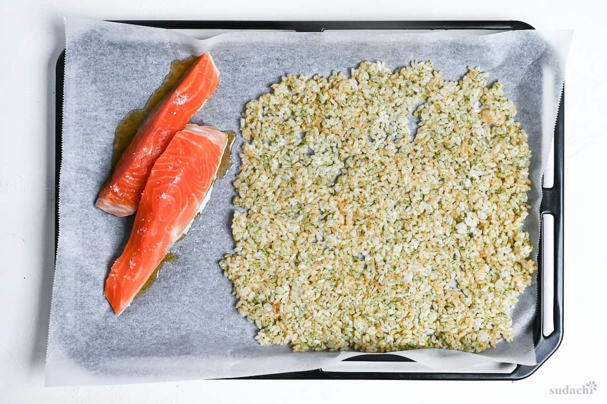 rice flattened into a single layer on an oven baking tray next to two raw salmon fillets