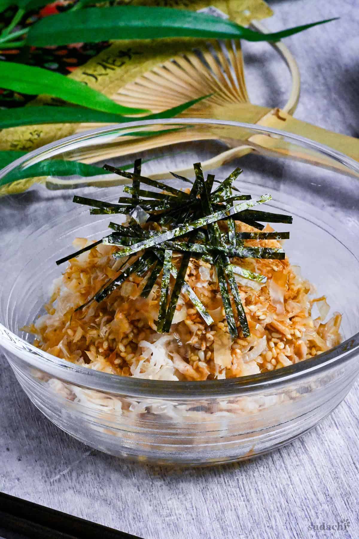 Addictive daikon radish salad topped with bonito flakes, sesame seeds and kizami nori served in a glass bowl next to black chopsticks and a Japanese fan in the background