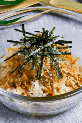 Addictive daikon radish salad topped with bonito flakes, sesame seeds and kizami nori served in a glass bowl next to black chopsticks and a Japanese fan in the background