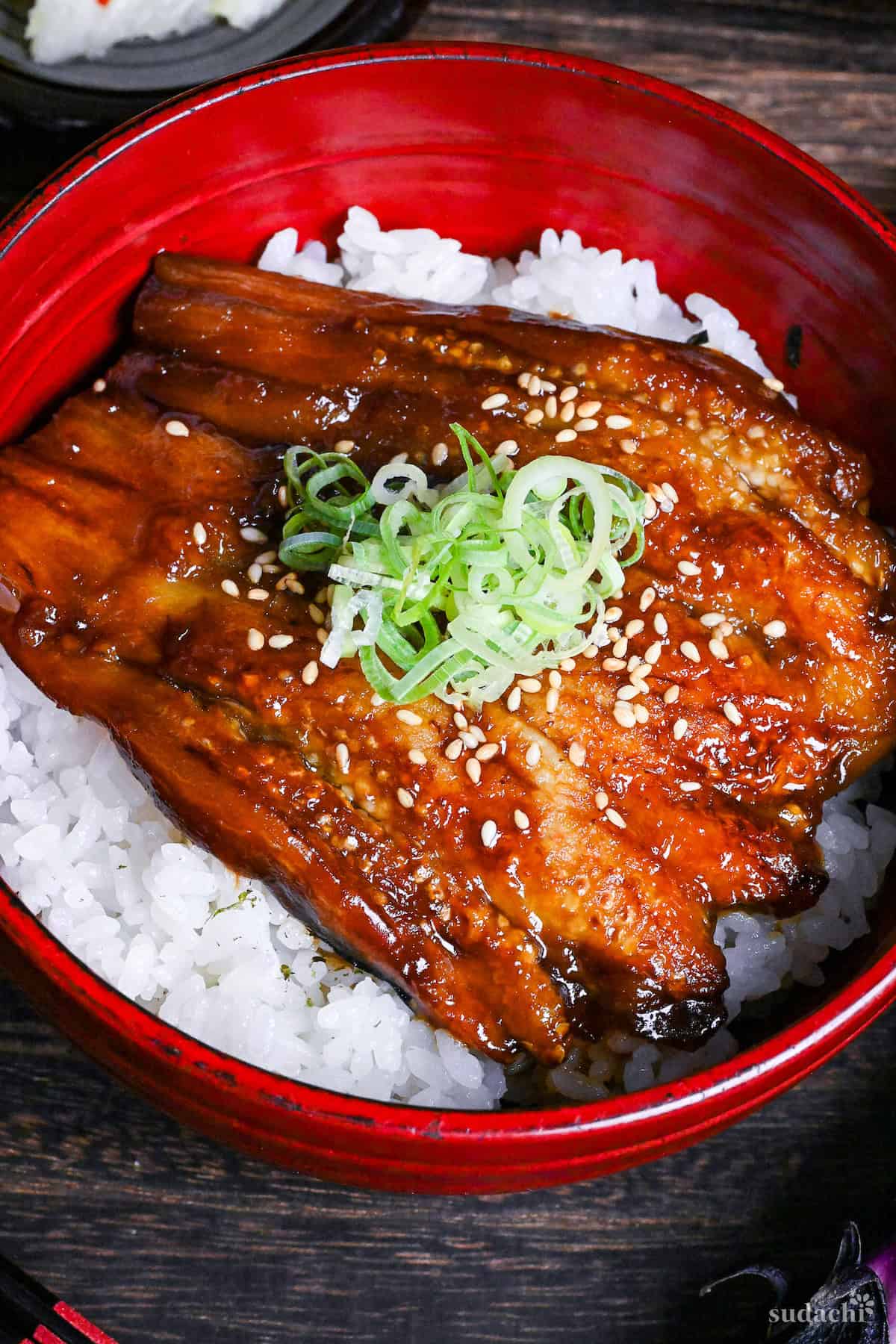 eel kabayaki in a red bowl on a dark wooden surface surrounded by raw eggplants, napa cabbage picjles and clear soup