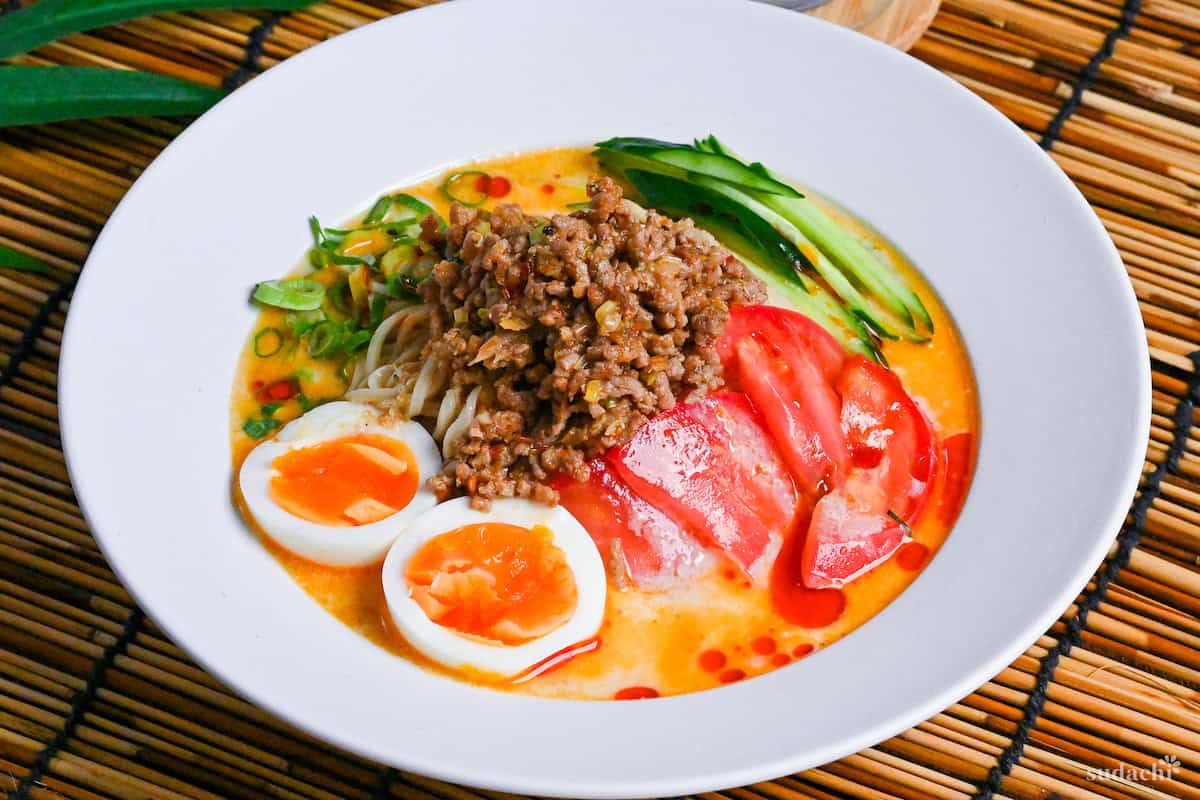 Hiyashi tantanmen in a white dish topped with boiled egg halves, julienned cucumber, chopped green onion, sliced tomato and seasoned ground pork on a bamboo mat background next to a glass of iced water and light wooden chopsticks side view