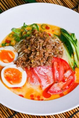 Hiyashi tantanmen in a white dish topped with boiled egg halves, julienned cucumber, chopped green onion, sliced tomato and seasoned ground pork on a bamboo mat background next to a glass of iced water and light wooden chopsticks