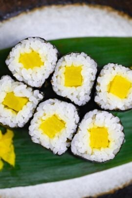 Six pieces of shinkomaki (pickled daikon sushi rolls) on a decorative leaf on a gray oval plate with dark brown rim