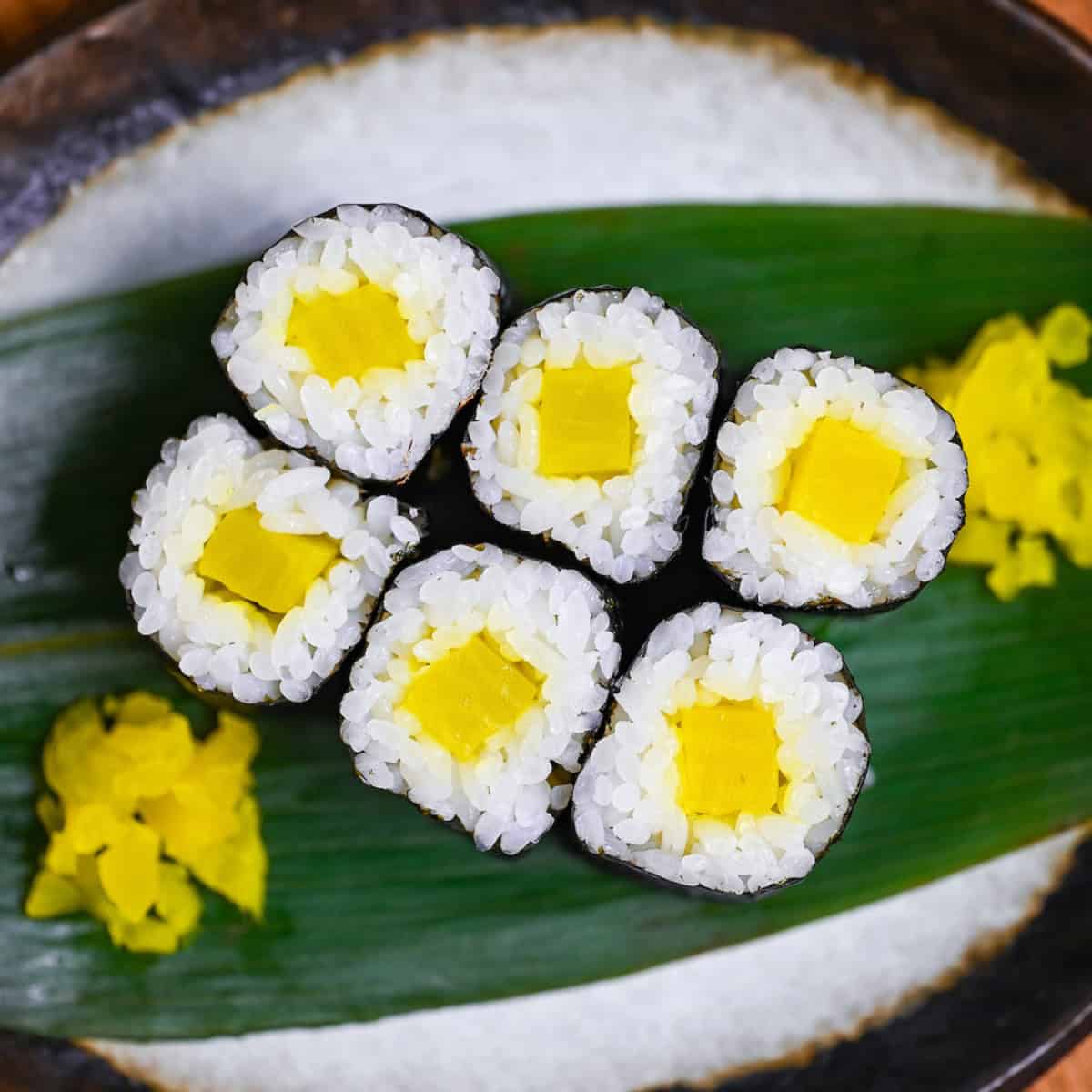 Six small sushi rolls filled with yellow daikon pickles on a leaf on a gray plate with brown rim