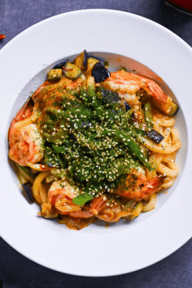 Spicy seafood yaki udon sprinkled with aonori and sesame seeds in a white dish on a gray surface surrounded by dry chilis and a jar of chili oil