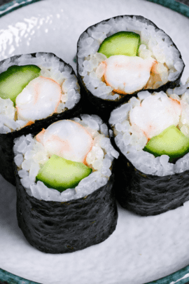 Four pieces of ebikyu maki made with cucumber and boiled shrimp coated with wasabi mayo on a white plate with teal rim on a gray background next to a fan and rice paddle