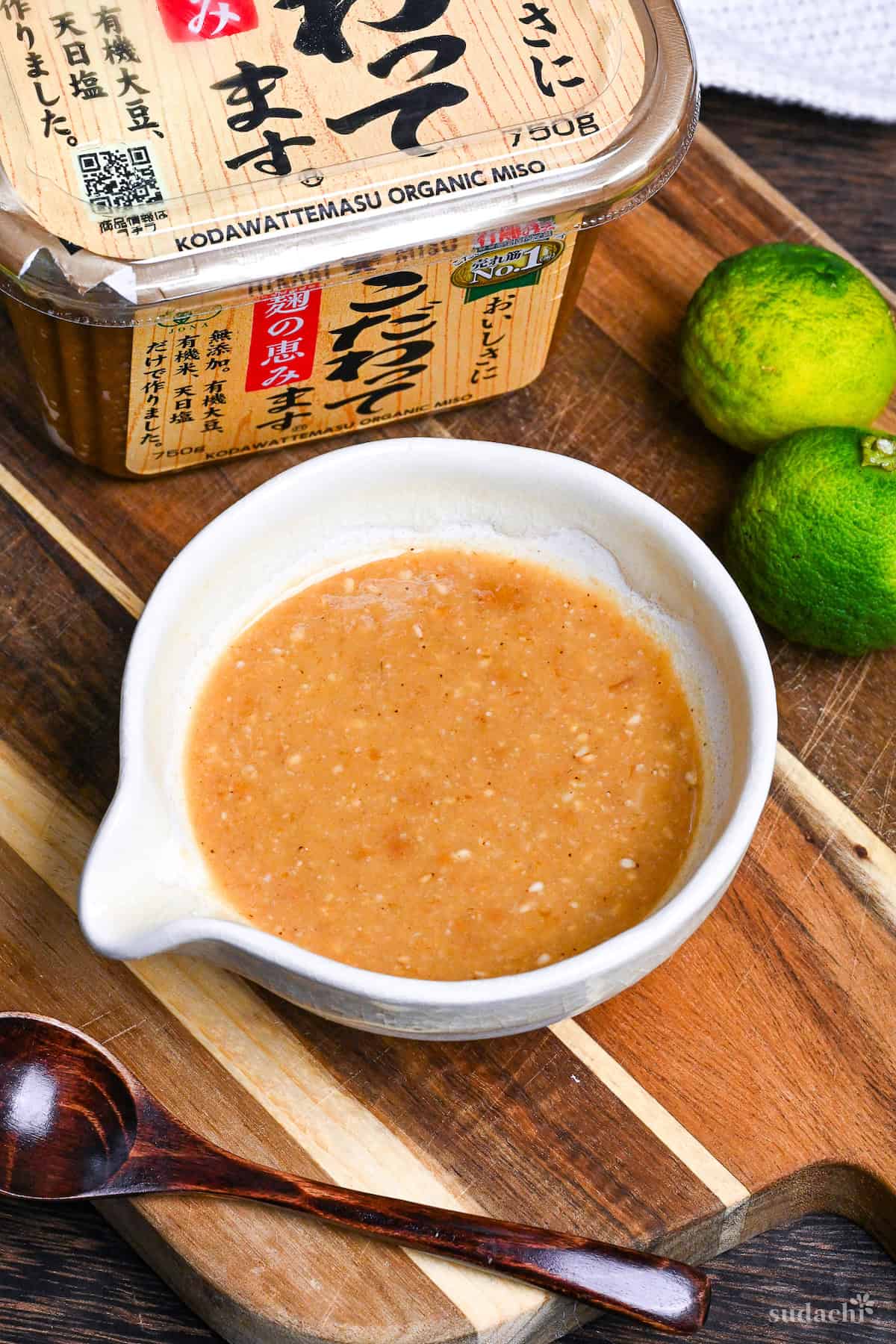 Homemade miso dressing in a Japanese style ceramic jug on a wooden chopping board infront a packet of yellow miso and two green yuzu