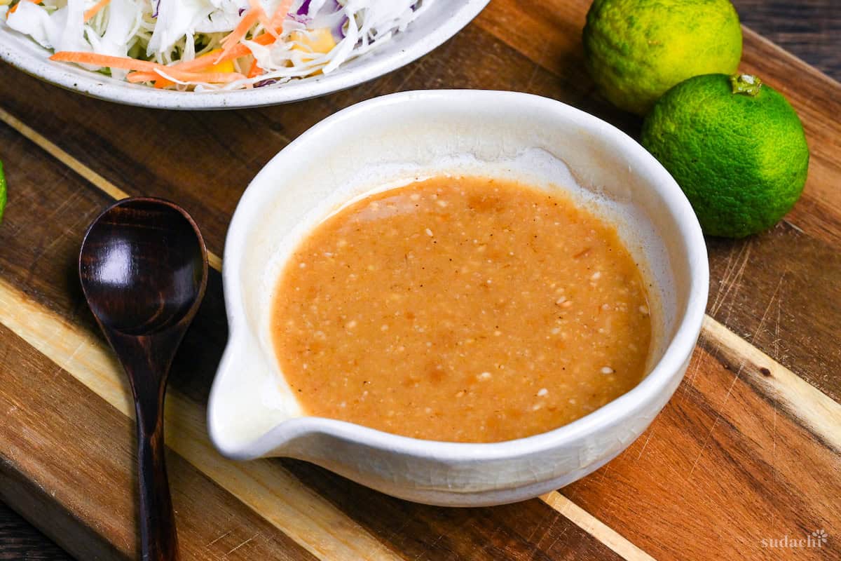 Homemade miso dressing in a Japanese style ceramic jug on a wooden chopping board next to a bowl of shredded vegetables and two green yuzu side view