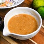 Homemade miso dressing in a Japanese style ceramic jug on a wooden chopping board next to a bowl of shredded vegetables and two green yuzu