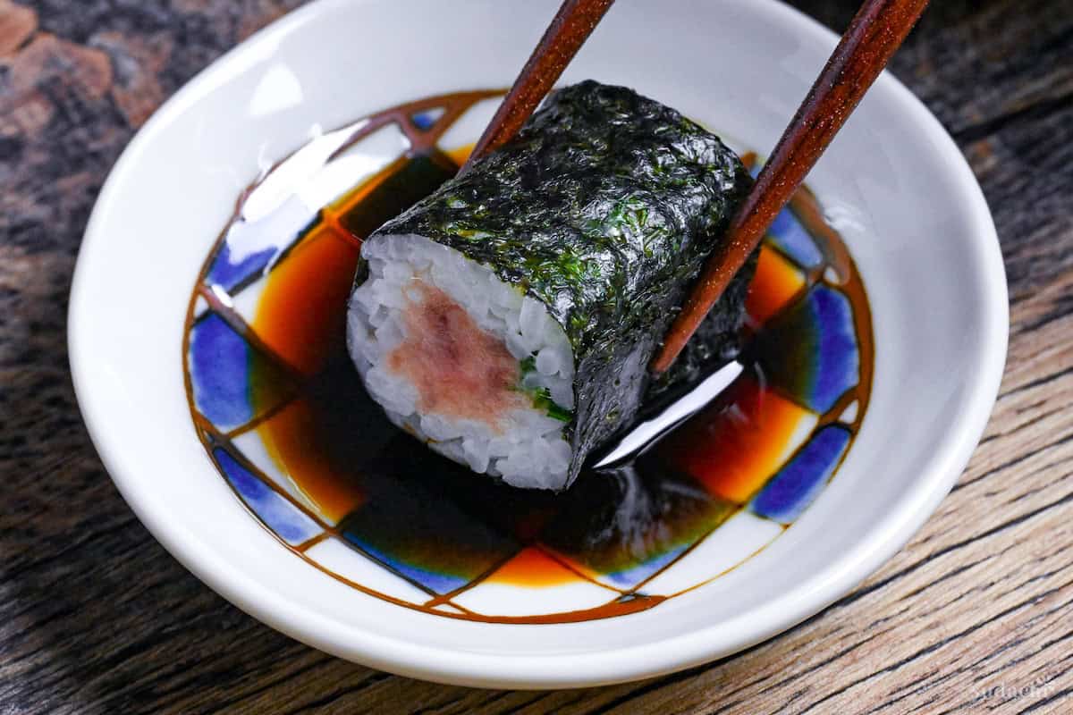 negitoro maki sushi roll held with wooden chopsticks dipped into a checkered bowl of soy sauce