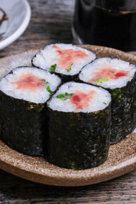 Four pieces of homemade negitoro sushi rolls on a brown serving dish with brush design next to a checkered dipping bowl and glass bottle of soy sauce