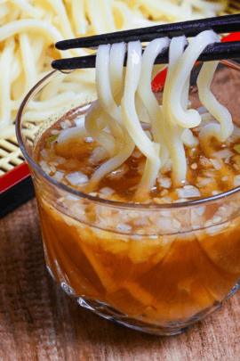 dipping ramen noodles in glass of chilled zaru ramen dipping sauce with cooked and chilled ramen noodles in the background with chashu and boiled eggs