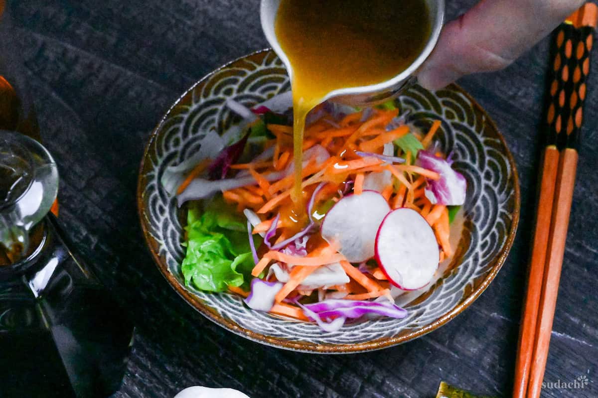 Japanese garlic dressing being poured over a colorful salad made with carrots, red cabbage, lettuce and radish in a brown and blue patterned Japanese bowl on a black background