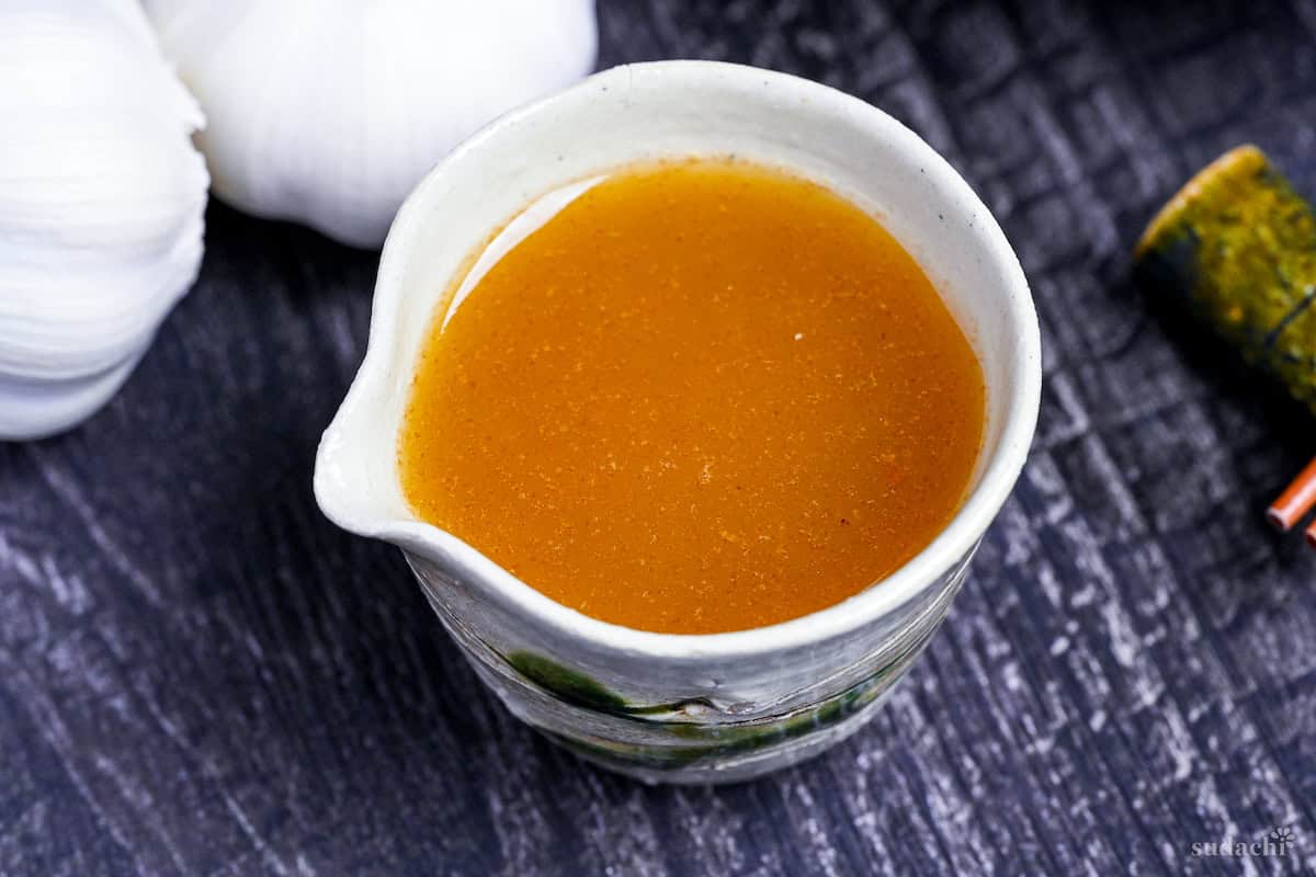 Japanese garlic dressing in a cream and green ceramic jug with garlic bulbs in the background on a black wood-effect background