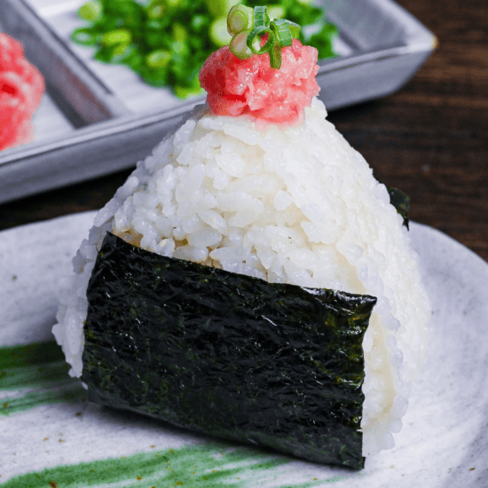 Negitoro onigiri (rice ball) on a gray plate with green brushstroke design, with mashed tuna and chopped green onions in a rectangular plate the background