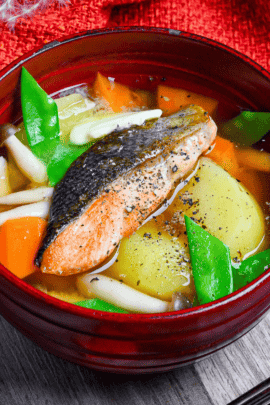 Salmon miso soup served in a dark red bowl on a gray wooden background with red cloth in the background