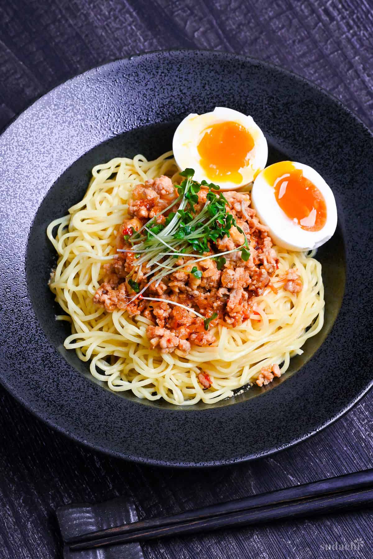 Shirunashi tantanmen topped with soft boiled eggs and broccoli sprouts served in a black dish on a black wood effect background