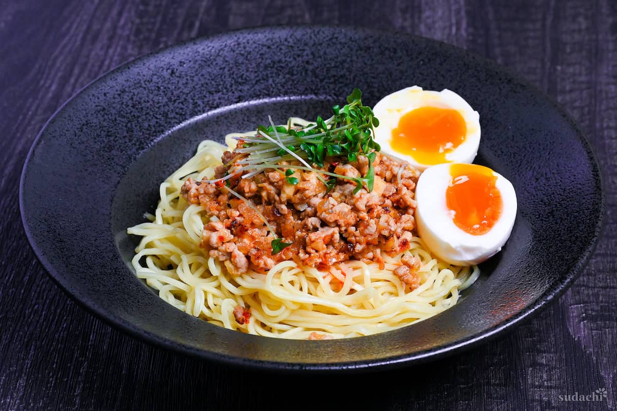 Shirunashi tantanmen topped with soft boiled eggs and broccoli sprouts served in a black dish on a black wood effect background side view