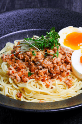 Shirunashi tantanmen topped with soft boiled eggs and broccoli sprouts served in a black dish on a black wood effect background