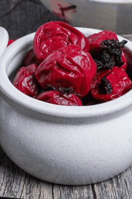 Homemade red umeboshi (Japanese pickled plums) in an off-white ceramic jar with lid set to the side on an ashy wooden surface