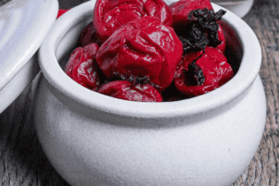 Homemade red umeboshi (Japanese pickled plums) in an off-white ceramic jar with lid set to the side on an ashy wooden surface