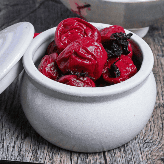 Homemade red umeboshi (Japanese pickled plums) in an off-white ceramic jar with lid set to the side on an ashy wooden surface