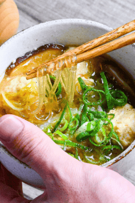 A hand holding harusame soup with chicken meatballs in a cream bowl holding up the glass noodles with wooden chopsticks with a wooden pepper grinder in the background