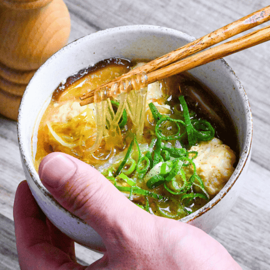 A hand holding harusame soup with chicken meatballs in a cream bowl holding up the glass noodles with wooden chopsticks with a wooden pepper grinder in the background