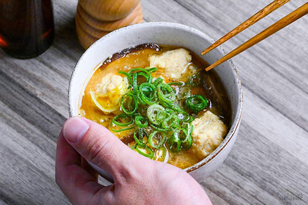 A hand holding a bowl of harusame soup with chicken meatballs topped with green onions and ground black pepper