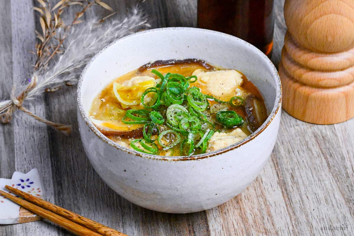 harusame soup with chicken meatballs in a cream bowl with a bottle of sesame oil and a wooden pepper grinder in the background