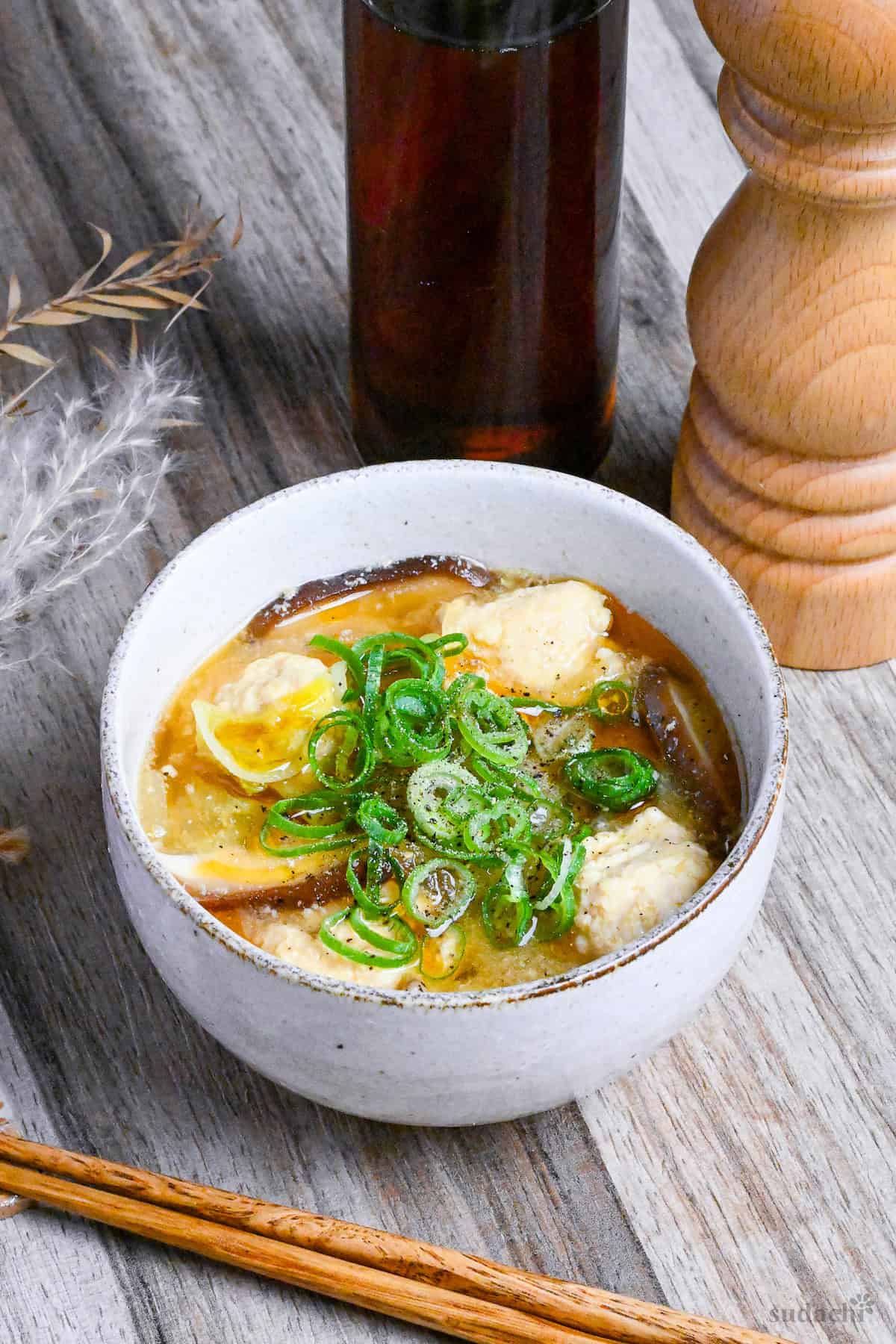 Harosami soup with chicken meat balls in a cream bowl with a bottle of sesame oil and wooden pepper in the background