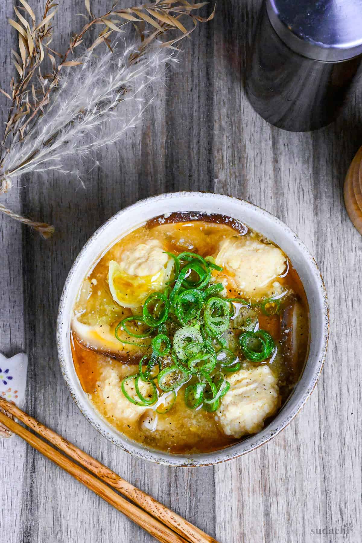 harusame soup with chicken meatballs in a cream bowl with a bottle of sesame oil and a wooden pepper grinder in the background top down view